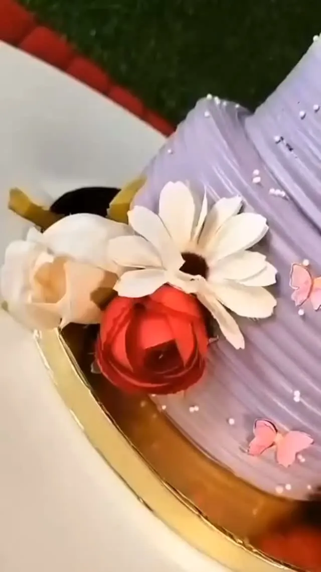 Close view of a lilac buttercream cake with combed ridges, a cream daisy, a deep red sculpted rose and small pink sugar butterflies on a gold cake board.