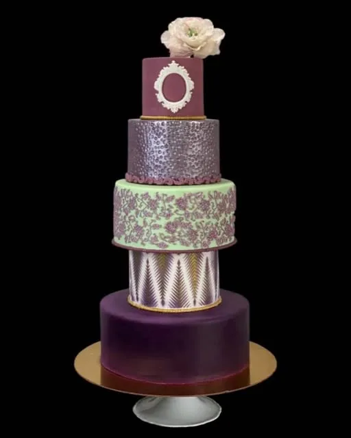 Five-tier celebration cake on a gold stand with plum, silver leaf, green lace-printed and metallic pleated tiers, topped with a single cream sugar flower.