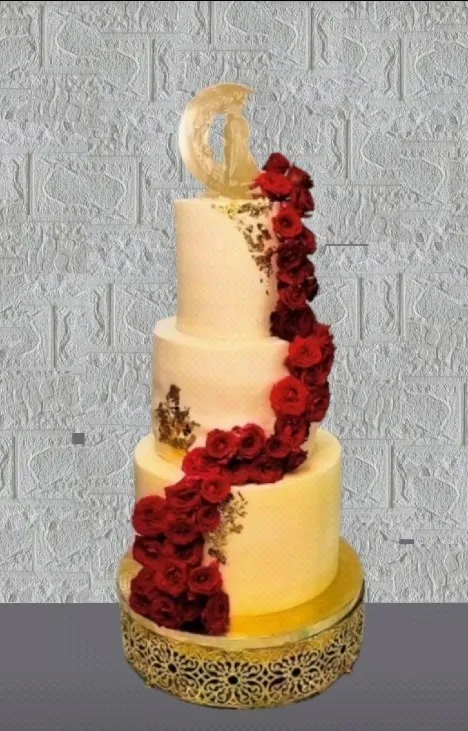 Three-tier ivory wedding cake decorated with a cascading trail of deep red roses, gold leaf accents and a gold monogram topper, on an ornate gold stand.